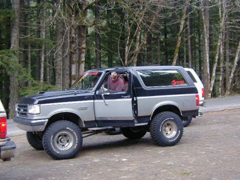 1994 Ford Explorer - A lifted Ford Bronco with off-road tires is parked on a dirt road, with a white SUV behind it in a wooded area.