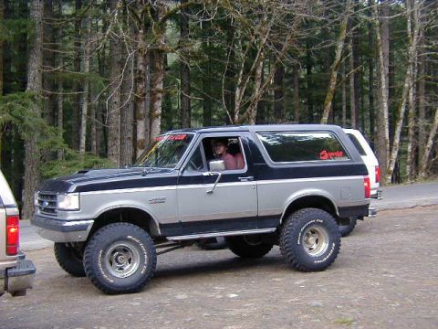 1994 Ford Explorer - A lifted, two-tone Ford Bronco is parked on a dirt road with trees in the background.