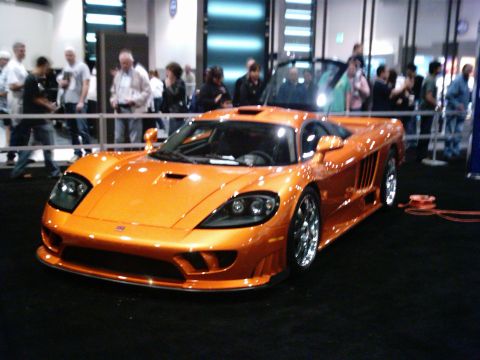 1986 Ford Bronco - A metallic orange mid-engine supercar on an auto show floor, shot from the front three-quarter angle with its butterfly door raised and hoo…