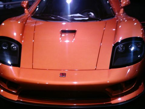 1986 Ford Bronco - A tight head-on shot of an orange Saleen S7 supercar under show lighting, showing the center hood vent, smoked quad-projector headlights, a…