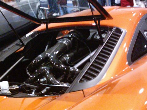 1986 Ford Bronco - Close-up of the open mid-engine bay of an orange supercar at an indoor show, showing gloss-black intake plumbing and tubular headers behind…