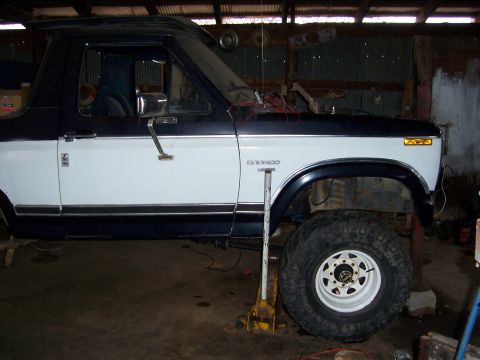 1983 Ford Bronco - This two-tone Ford Bronco is shown in profile, lifted by a yellow jack, with its front wheel visible.