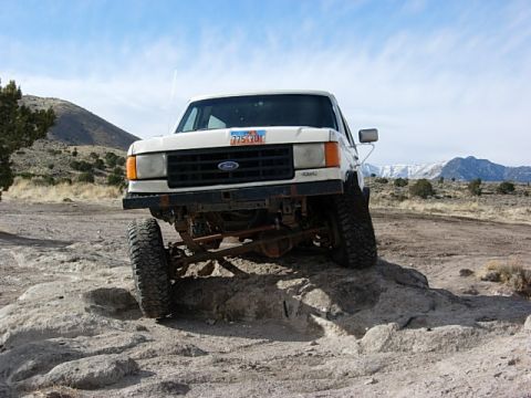 1987 Ford Bronco - This lifted Ford F-Series truck is navigating a rocky trail under a partly cloudy sky.