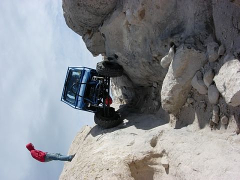 1987 Ford Bronco - A person in a red jacket watches as a blue off-road vehicle teeters on the edge of a steep cliff.