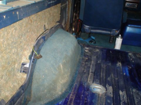 1984 Ford Bronco - The rear interior of a vehicle shows blue seats and carpet, with exposed insulation on the side panel.