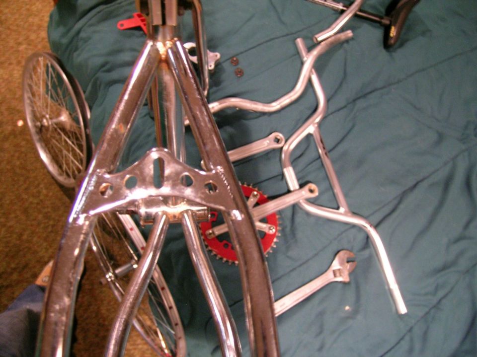 Disassembled chrome bicycle parts including fork, wheel, handlebars and red chainring on a green blanket