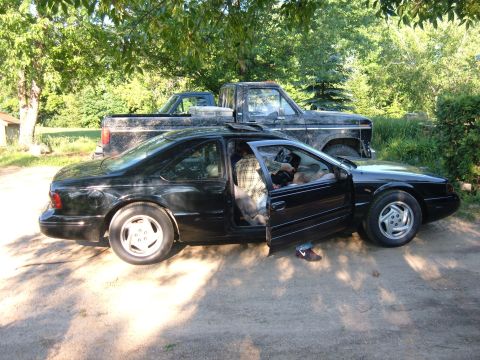 1985 Ford F-150 - Two vehicles, a black coupe and a black pickup truck, are parked outdoors on a dirt surface.