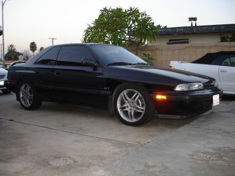 1989 Mazda MX-6 - A black coupe with tinted windows and custom wheels is parked on a concrete surface next to a white convertible.