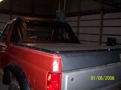 1985 Ford Bronco - This red SUV has a black soft top and a grey tailgate, viewed from the rear in a garage.