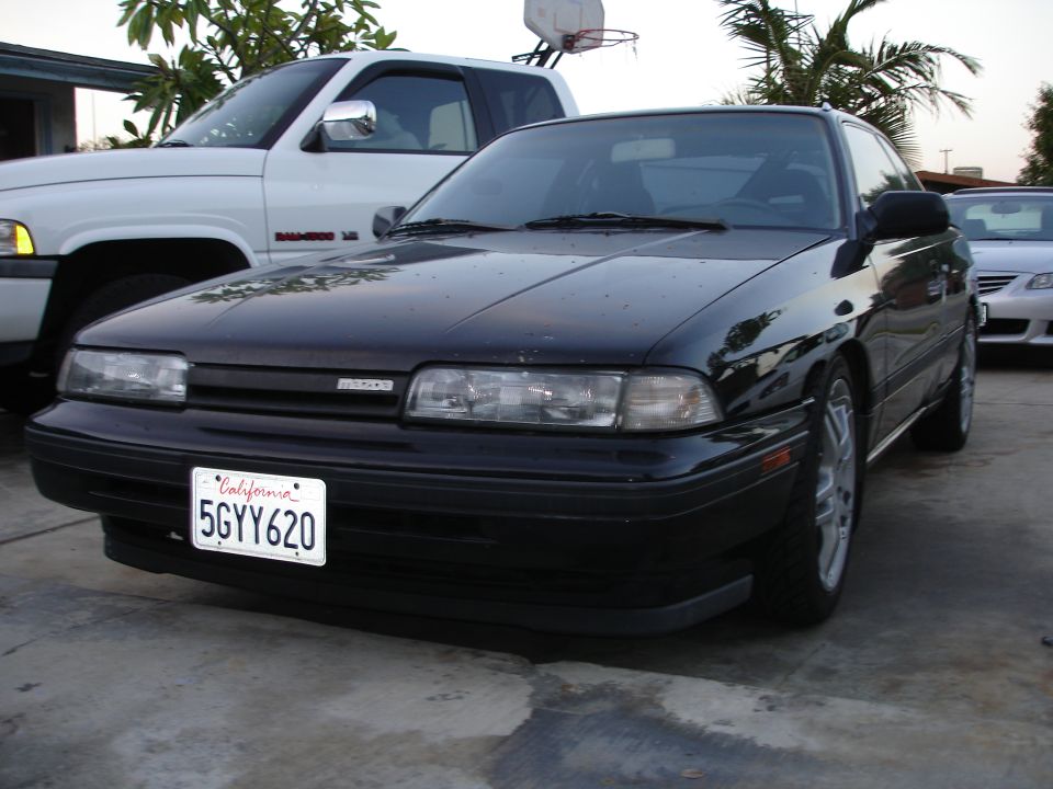 Black 1989 Mazda MX-6 coupe parked next to a white pickup truck