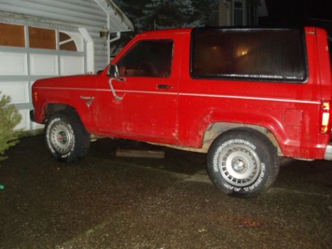 1985 Ford Bronco II - A red 1985 Ford Bronco II is parked on a wet driveway next to a garage.