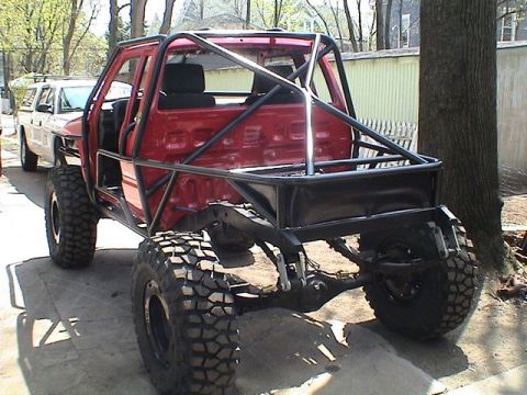 1986 Ford F-150 - This heavily modified red pickup truck features a custom roll cage and oversized tires, parked next to a white truck.