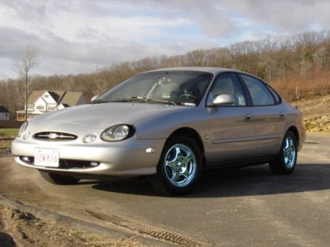 1999 Ford Taurus - Silver 1999 Ford Taurus sedan parked on a paved surface with trees and houses in the background.