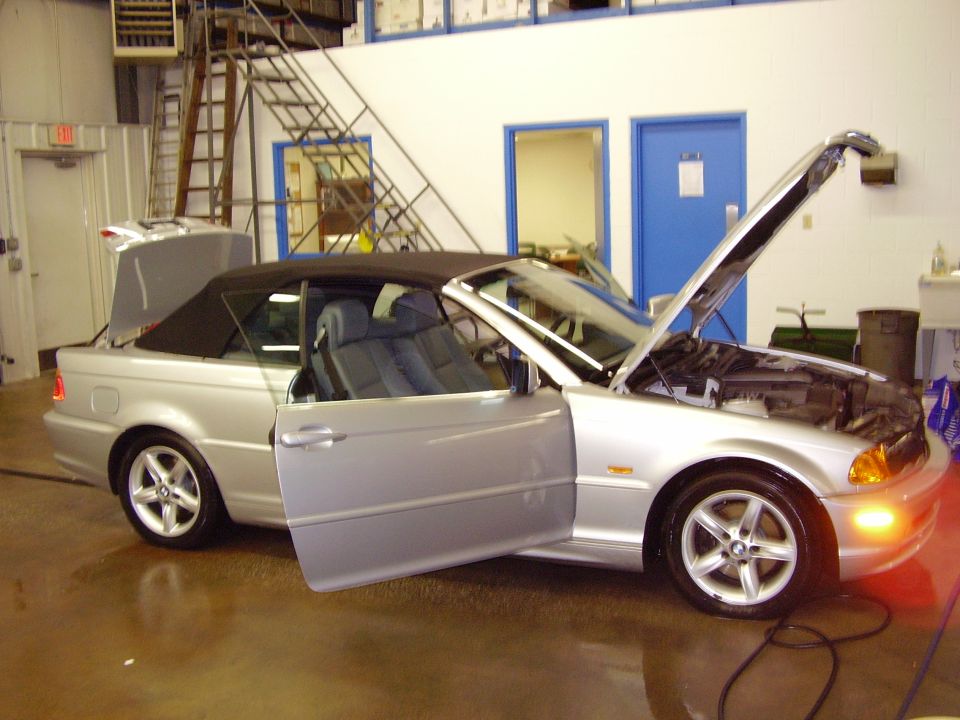 Silver convertible with driver's door, trunk, and hood open. Interior shows blue seats
