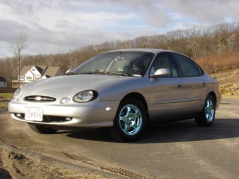 1999 Ford Taurus - This 1999 Ford Taurus is captured in a three-quarter front view, its silver exterior against a backdrop of trees and houses.