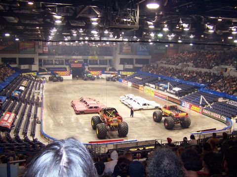 1995 Nissan Frontier - Monster trucks are prepared for an event in an arena, with crushed cars on the floor.