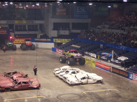 1995 Nissan Frontier - Monster trucks are set up in an arena with crushed cars as obstacles, with spectators in the stands.