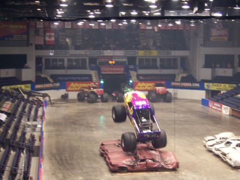 1995 Nissan Frontier - Monster trucks perform in an arena, with one truck airborne over a pile of crushed cars.