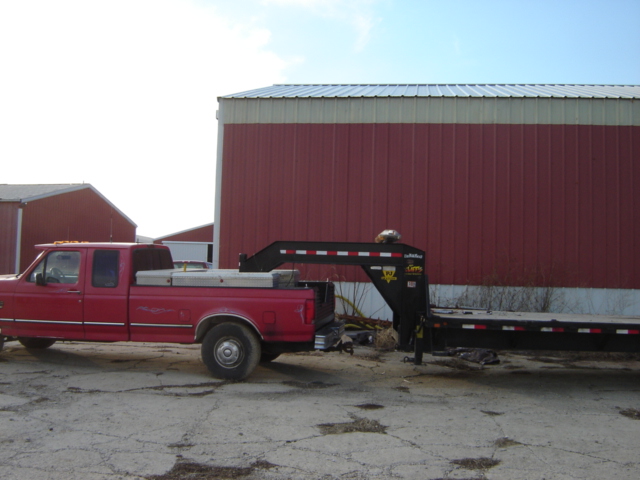 Red pickup truck with a toolbox in the bed, hitched to a black gooseneck trailer. Large red barn in background