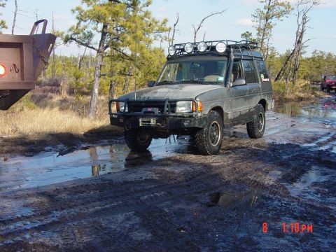 1995 Dodge 1500 - An off-road vehicle navigates a muddy trail with trees and water in the background.
