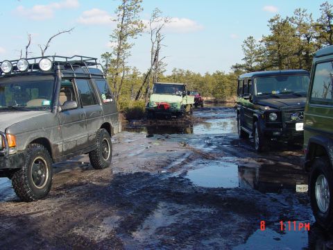 1995 Dodge 1500 - A group of off-road vehicles traverses a challenging muddy terrain, surrounded by trees and water.