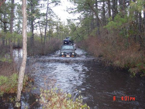 1995 Dodge 1500 - Two off-road vehicles navigate a water-filled path surrounded by trees and brush.