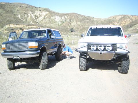 1990 Ford Bronco - Two off-road modified trucks, a dark blue Bronco and a white pickup, are parked on a dirt road with mountains in the background.