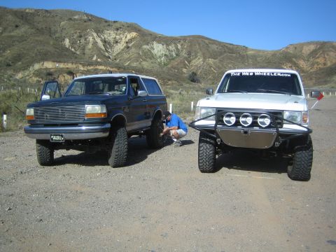 1990 Ford Bronco - blue SUV and a white SUV with a brush guard park side by side on dirt while a person kneels between them in dry hills.