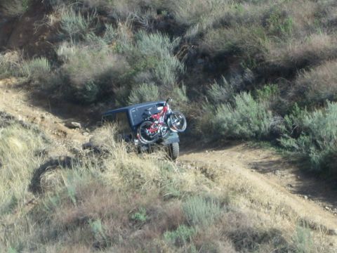 1990 Ford Bronco - dark off-road vehicle carrying bicycles on a rear rack descends a steep dirt trail through brushy hills.