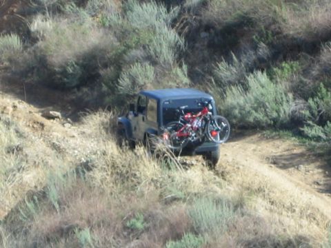 1990 Ford Bronco - blue Jeep Wrangler with a hardtop and rear-mounted bike rack drives a dirt trail through scrubby hills.