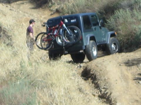 1990 Ford Bronco - gray Jeep Wrangler carries two mountain bikes on a rear rack as a man stands beside it on a rutted dirt trail.
