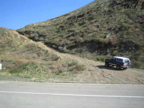 1990 Ford Bronco - dark full-size SUV is parked on a dirt shoulder beside a paved road at the foot of a dry grassy hillside.
