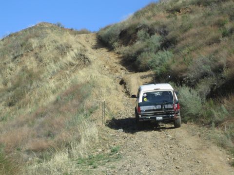 1990 Ford Bronco - white SUV with a roof rack climbs a steep dirt trail up a dry grassy hillside under a blue sky.