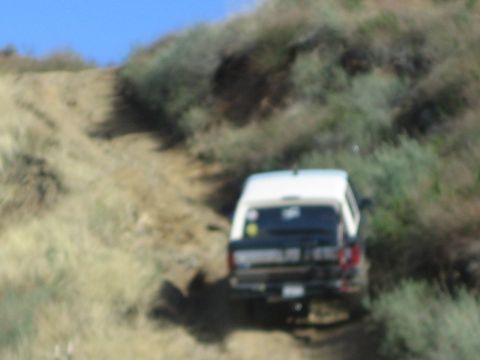 1990 Ford Bronco - white-topped, dark-bodied SUV with a rear tire carrier climbs a steep, rutted dirt trail among brush; slightly blurred.