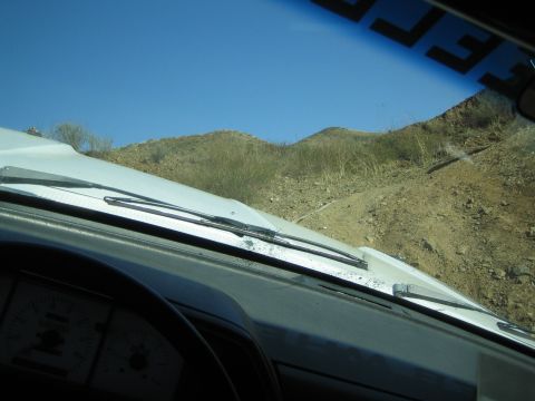 1990 Ford Bronco - From inside a vehicle, the view over a white hood and dashboard gauges looks out to a dirt trail and dry hills.