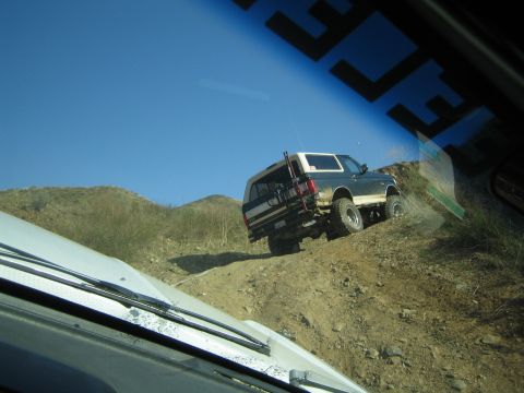 1990 Ford Bronco - Seen through a windshield, a lifted dark SUV with a white top and rear tire carrier climbs a rocky dirt trail on a dry hill.