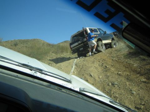 1990 Ford Bronco - Through a windshield, a man in a blue shirt stands next to a lifted dark-and-white SUV stopped on a steep dirt trail.