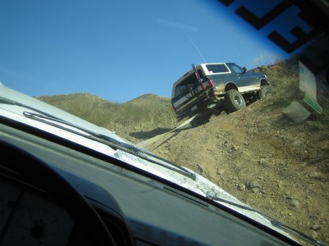 1990 Ford Bronco - Through a windshield over a white hood, a lifted SUV with a white top climbs a steep rocky dirt incline on a dry hillside.