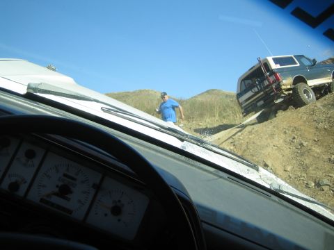 1990 Ford Bronco - Through a windshield showing dashboard gauges, a man in blue walks toward a lifted white-topped SUV on a steep dirt trail.