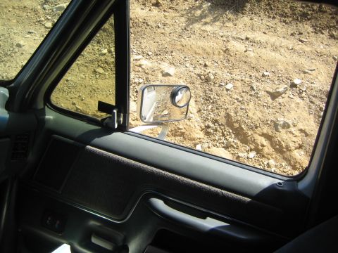 1990 Ford Bronco - View from inside an SUV looking out the passenger window past a clip-on towing mirror toward a steep dirt embankment.