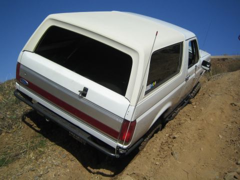 1990 Ford Bronco - white two-door Ford Bronco with a red side stripe sits at a steep angle on a dirt off-road trail under a blue sky.