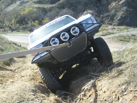 1990 Ford Bronco - white lifted truck with a tubular brush guard, three round off-road lights and large mud tires articulates over a rocky off-road obstacle.