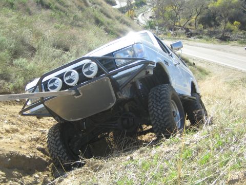 1990 Ford Bronco - white lifted truck with a tube brush guard, three round lights and big mud tires leans steeply on a dirt embankment beside a paved road.