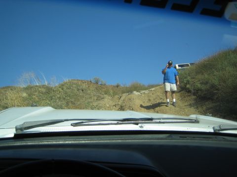 1990 Ford Bronco - Shot through a windshield over a white hood: a man in a blue shirt and tan shorts stands on a steep dirt trail under clear blue sky.