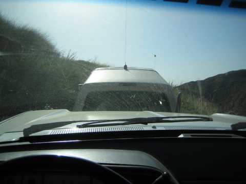 1990 Ford Bronco - Looking through a dusty windshield over the dashboard at a white SUV ahead on a dirt trail among brown hills.