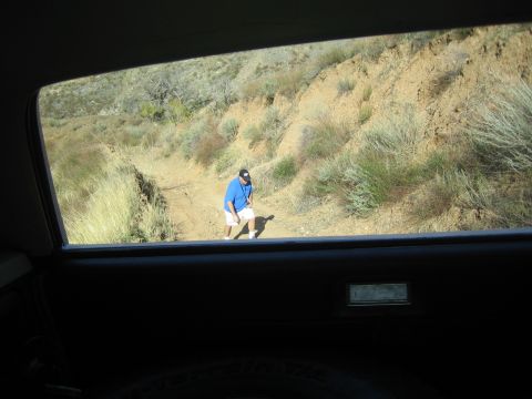 1990 Ford Bronco - Through a vehicle's rear side window, a man in a blue shirt walks along a dirt trail beside an eroded hillside.