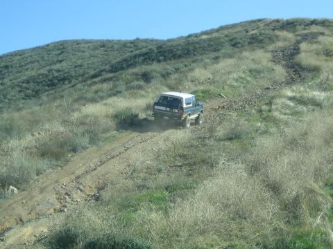 1990 Ford Bronco - dark and white two-tone SUV climbs a rutted dirt trail up a grassy hillside, kicking up dust under a clear sky.