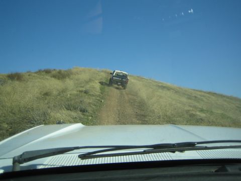 1990 Ford Bronco - Through a windshield over a white hood, a white SUV descends a steep grassy hill trail under a clear blue sky.