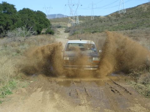 1990 Ford Bronco - truck blasts through a muddy puddle sending up large sprays of brown water on a dirt road beneath power transmission towers.