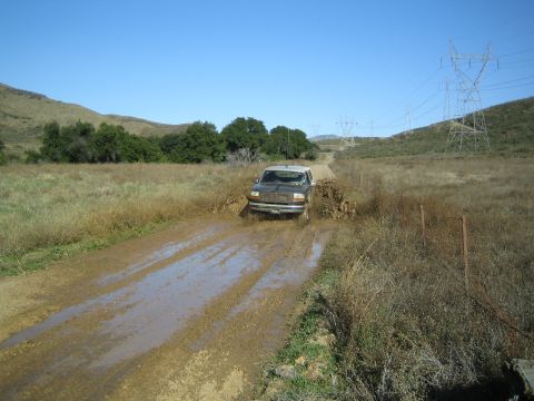 1990 Ford Bronco - mud-covered SUV drives through a flooded, muddy dirt road in a grassy valley lined with power transmission towers.
